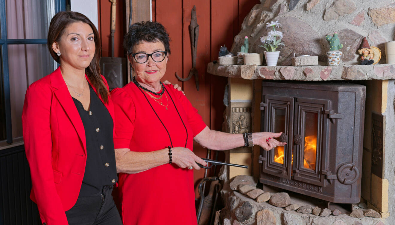 Ida och Irene står bredvid en öppen kakelugn, elden brinner och Irene står med en eldgaffel i ena handen och håller i spisluckan.