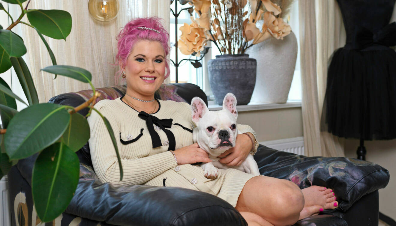 Porträttbild på Melinda Jacobs. Hon har rosa hår som är uppsatt. Hon sitter i en svart skinnfåtölj och håller i sin ljusa hund. Hon har på sig en beige stickad klänning.