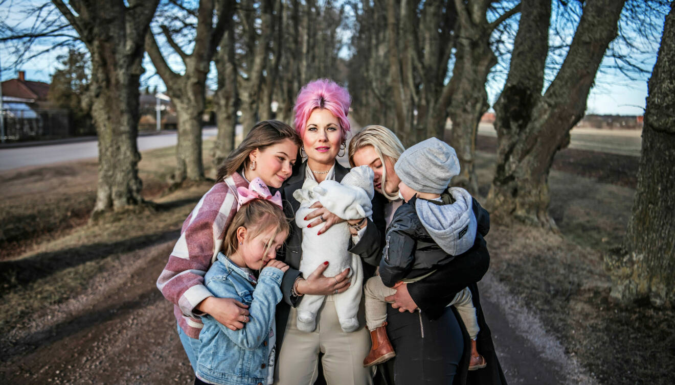 Melinda tillsammans med fem barn utomhus, i famnen håller hon en bebis i vit overall. I bakgrunden syns träd.