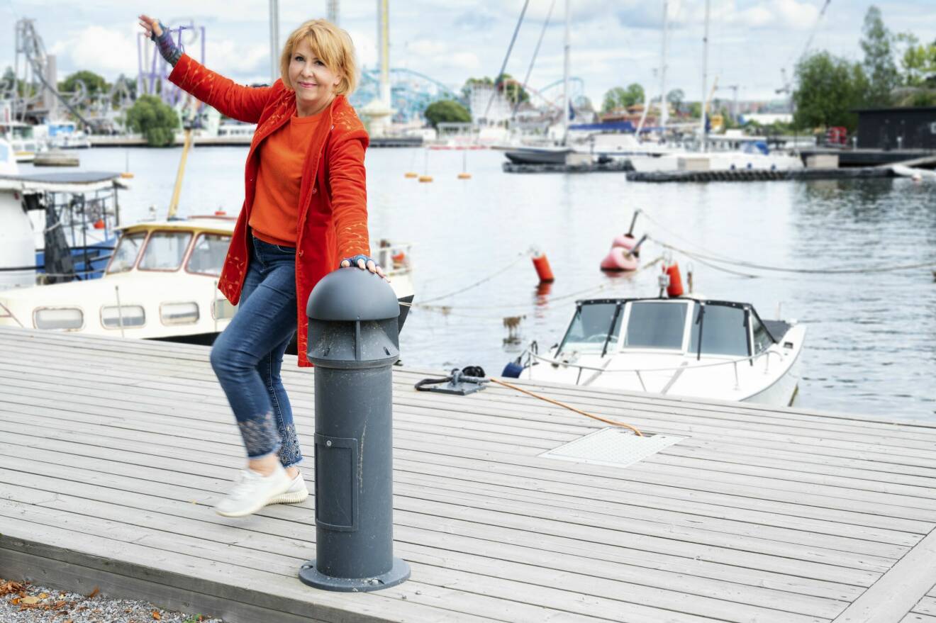 Porträttbild på Pia Johansson som har på sig en orange kavaj och tröja. Hon står på en båtbrygga, lutar sig mot en stolpe och höjer den ena handen ovanför huvudet. Hon ler och tittar in i kameran.
