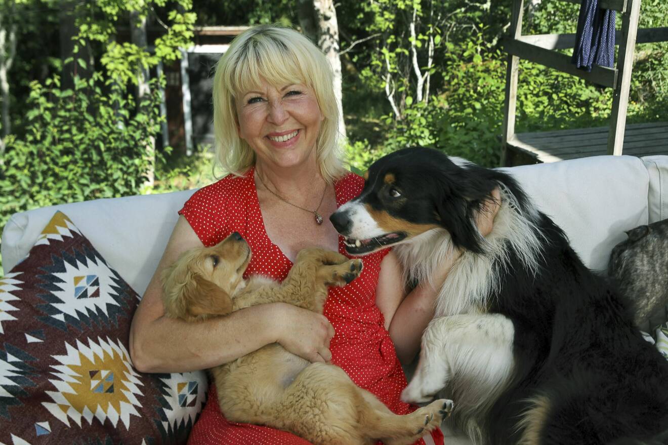 Bild på Pia, hon sitter i en soffa utomhus, med två hundar i knät. Hon ler och håller om hundarna. Hon har en röd topp på sig.