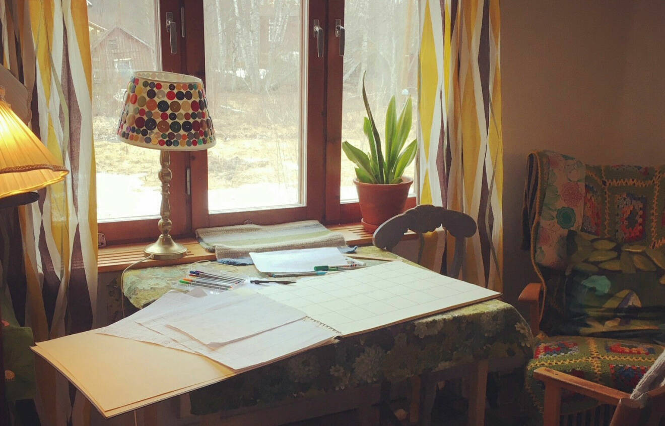 Ett skrivbord framför ett fönster. På skrivbordet ligger massa papper och pennor.