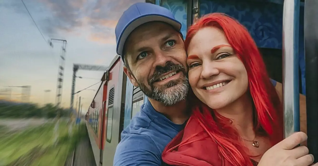Anders och Lousie lutar sig ut från ett tågfönster under en resa. Mannen har blå tröja och keps på sig. Kvinnan har rött hår, ögonbryn och tröja.