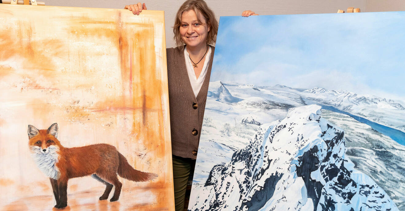 Karin står med två stora målningar som hon gjort, en på en räv och den andra på ett snötäckt berg.