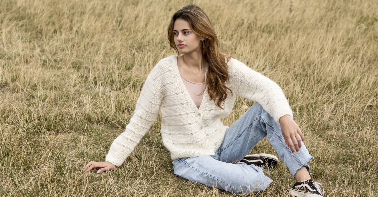 Ung kvinna i jeans och vit kofta sitter i gräset.