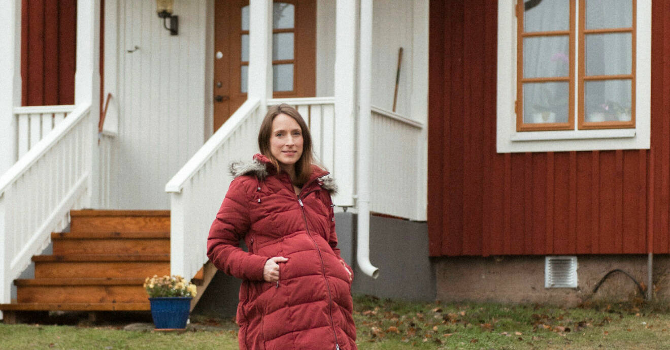 Jenni står framför sitt hus, hon har på sig en röd lång dunjacka och hon är gravid.