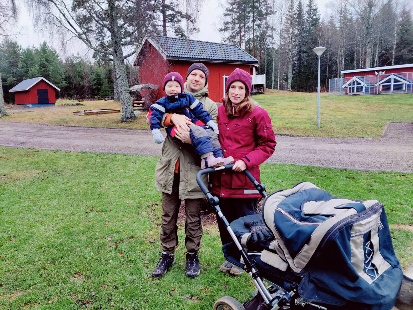 Jenni har på sig en röd jacka och mössa. Tillsammans med sin man Per som håller dottern Elin i famnen. De är utomhus och står vid en barnvagn.