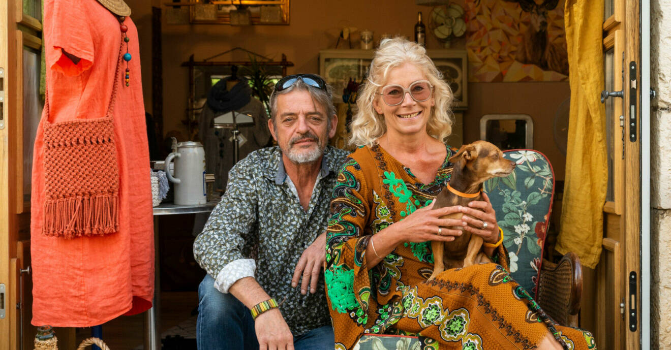 Fred och Sonia sitter utanför sin loppisbutik. Sonia håller sin hund i famnen och har en färgglad mönstrad klänning på sig.
