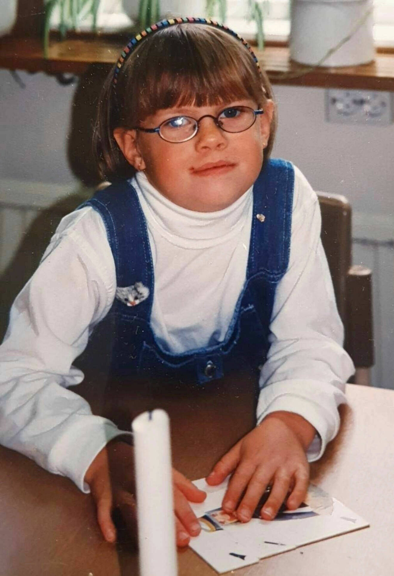 En ung flicka på 1990-talet, hon har glasögon, vit polotröja och blå klänning över.