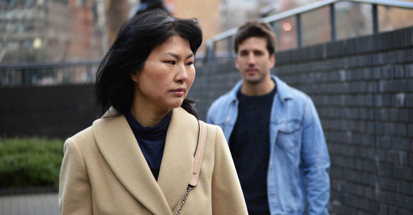 Kvinna har på sig en beige kappa och bakom henne står en man i blå jeansskjorta.