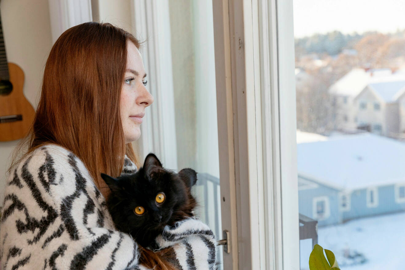 Elin har rött långt hår och tittar ut genom ett fönster. Hon har en svart katt i famnen och en zebra-mönstrad tröja.