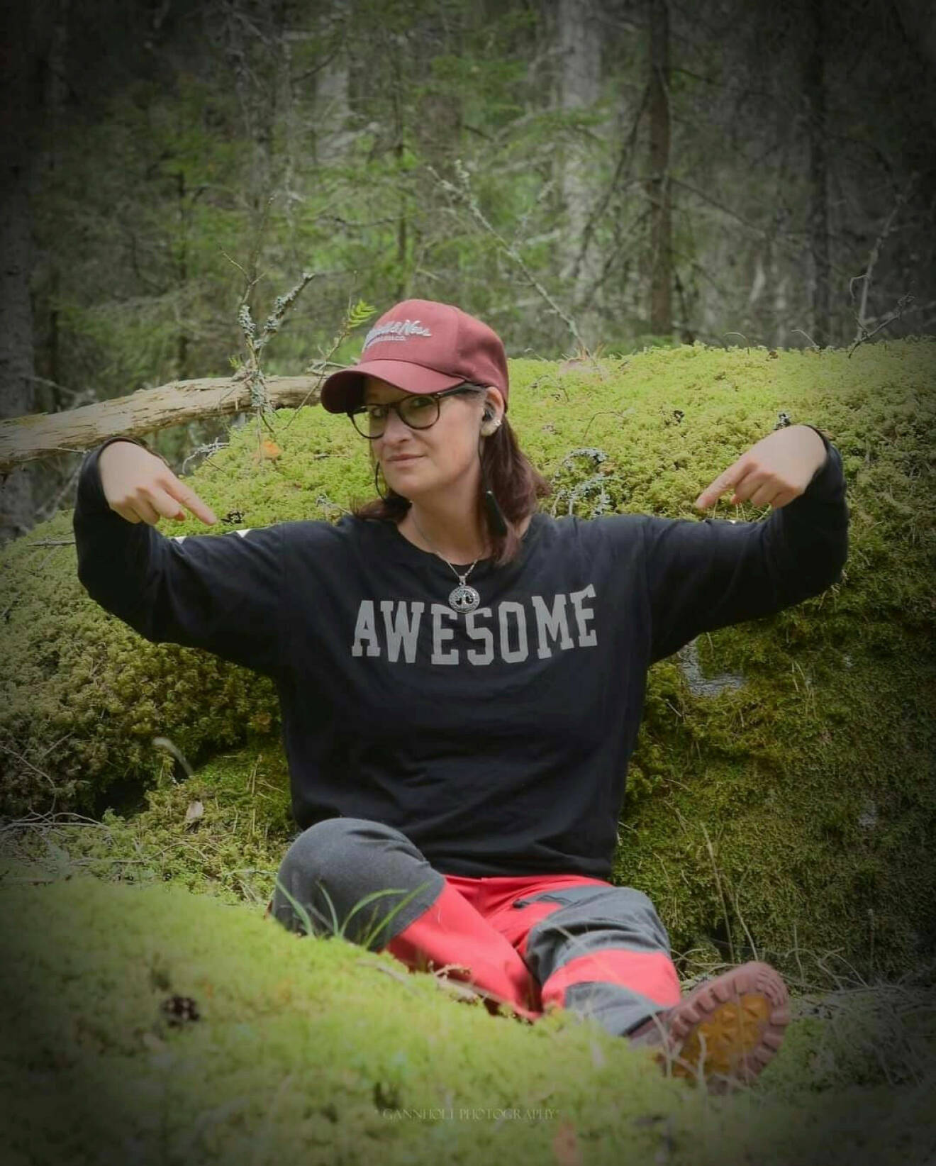 Kristina sitter bland mossan i skogen. Hon har på sig en keps och en tröja med orden "awesome".