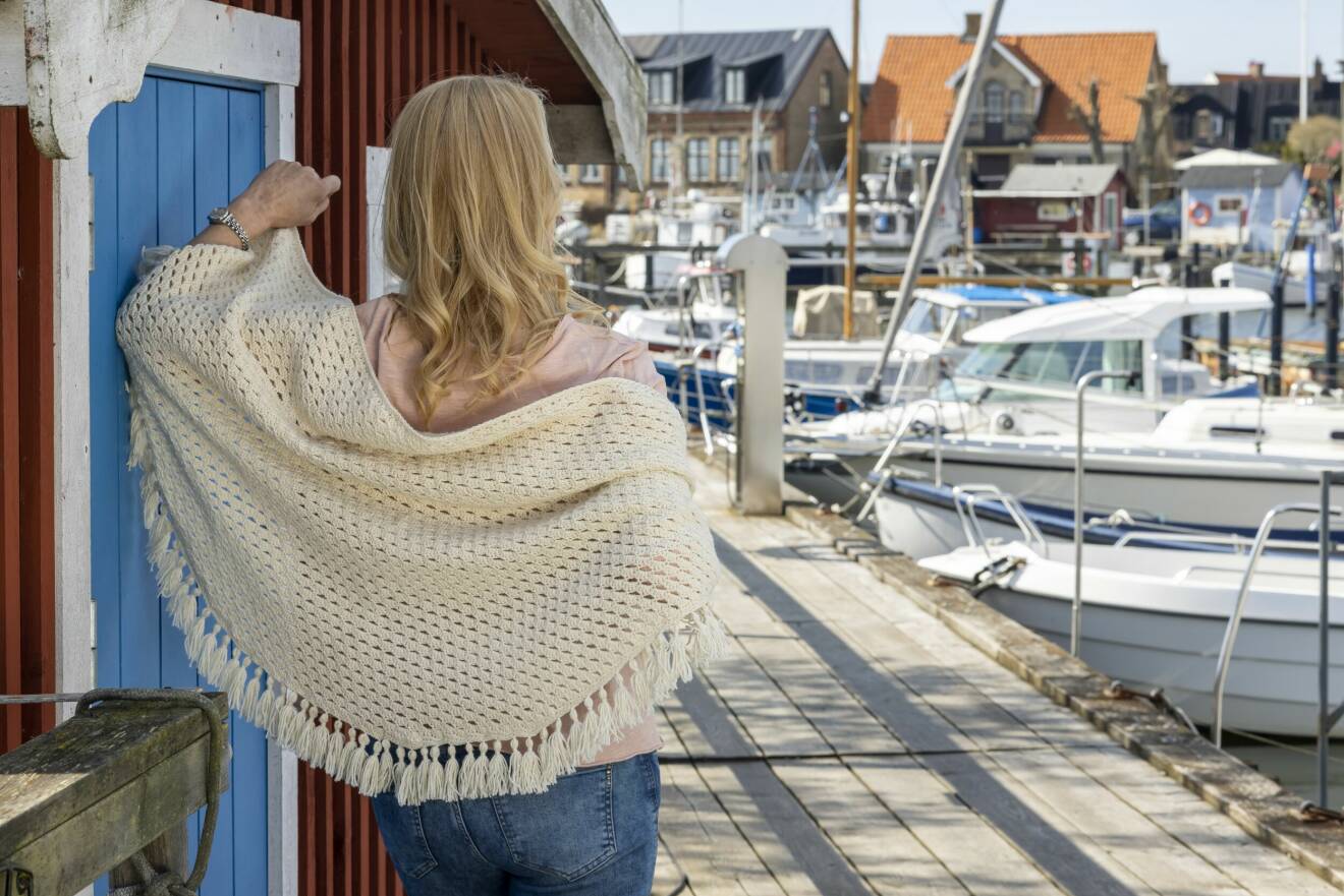 Kvinna har på sig en virkad vit sjal, hon står på en brygga vid en hamn.
