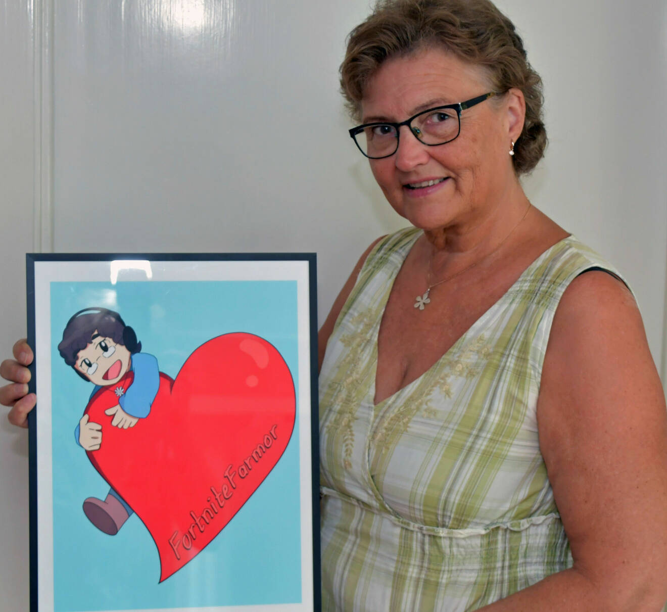 Susanne med en tavla som föreställer hennes egna emoji, en kvinna med glasögon, halsband som kramar ett rött hjärta i vilket det står FortniteFarmor.