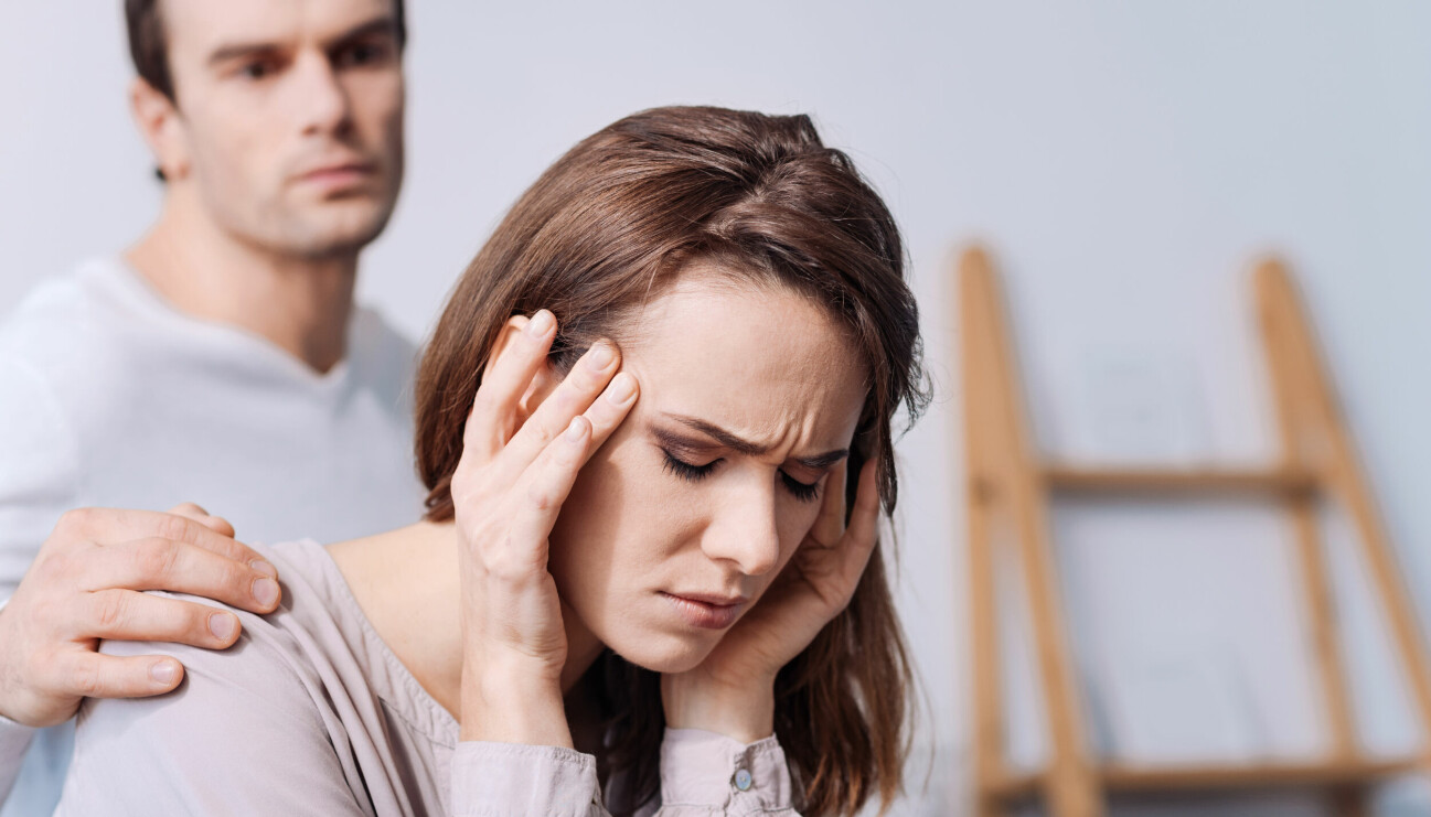 Ung kvinna med huvudvärk masserar sina tinningar, medan hennes pojkvän lägger en stöttande hand på hennes axel.