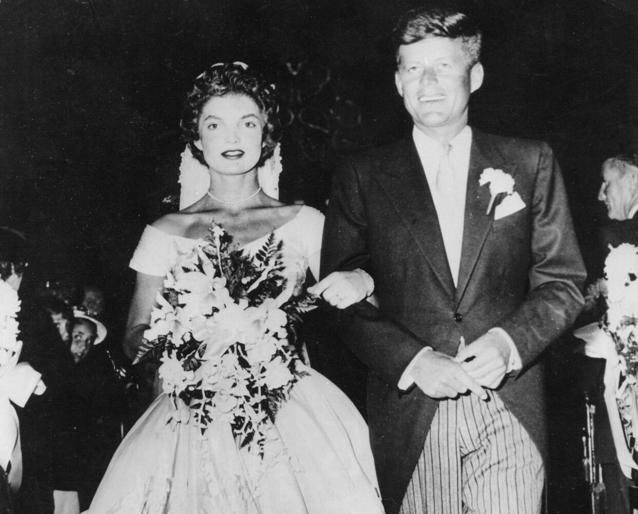 1953 gifte sig Jacqueline Bouvier med John F Kennedy. Jaqueline Bouvier bär vit bröllopsklänning, slöja och brudbukett och John F Kennedy bär frack och randiga byxor.
