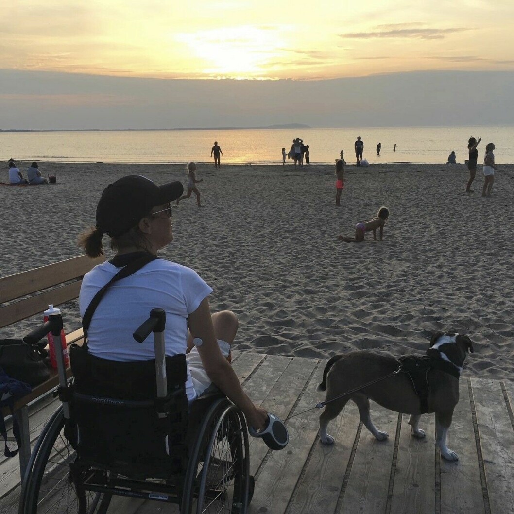 Jessica ser livet pågå på stranden nedanför henne. Vid den här tiden tampades hon mot såväl benbrott som tät dialys. Jessica Lindgren, under tiden hon tampas med ständig dialys samtidigt som hon sitter i rullstol på grund av benbrott. Här sitter hon i rullstolen vid havet, med hunden Elsa vid sin sida, och ser strandlivet pågå.