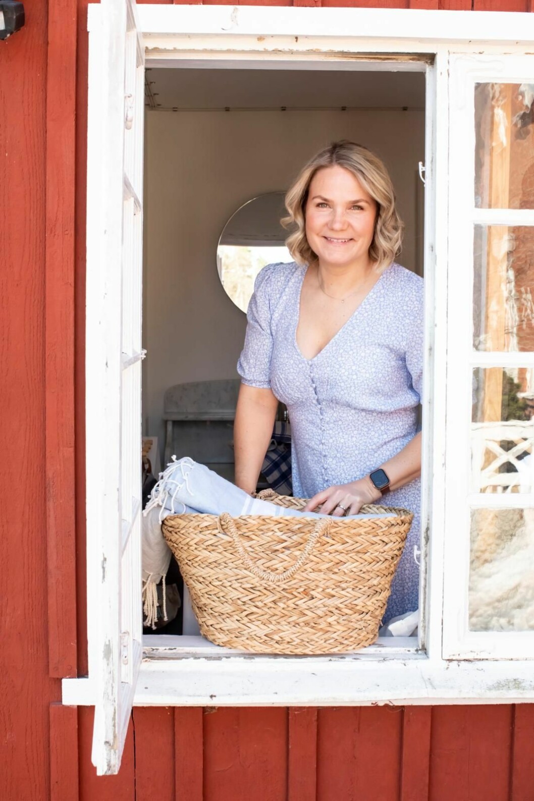 Johanna Bank tittar leende ut genom ett öppet fönster. Framför henne står en korg med blåvit hamamduk.