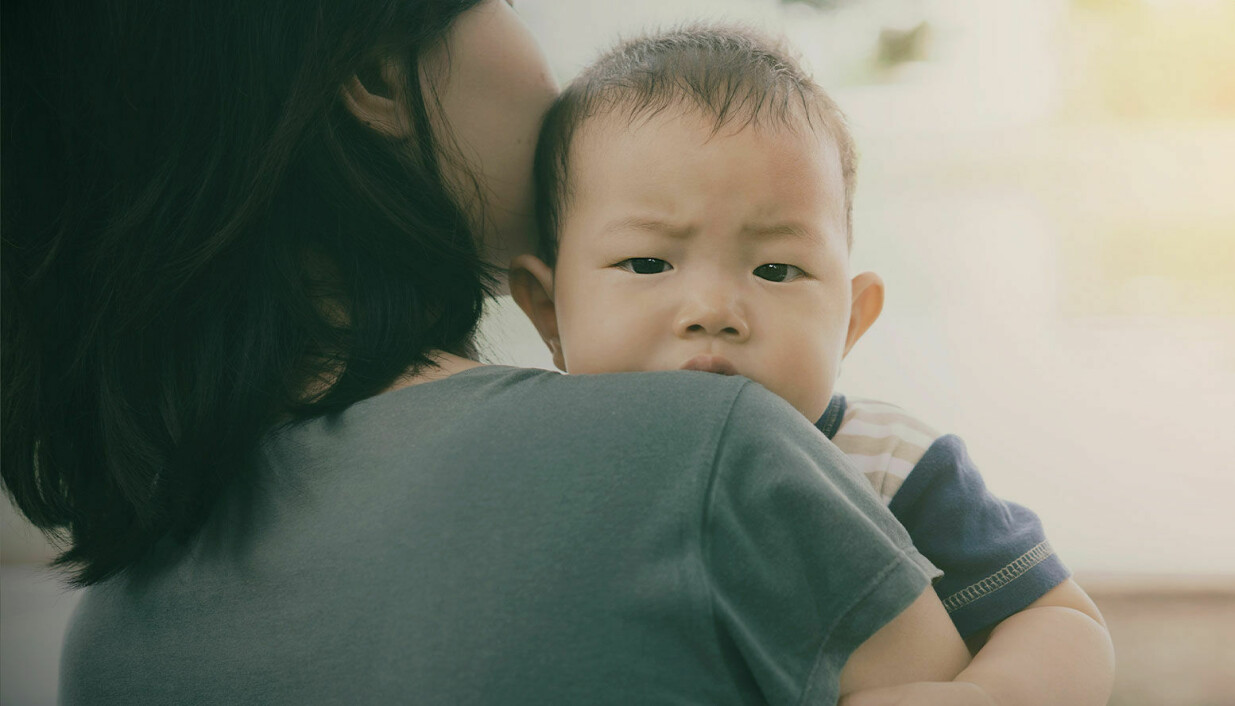 En kvinna står med ryggen mot kameran. I sin famn håller hon ett litet barn som tittar in i kameran över sin mammas axel.