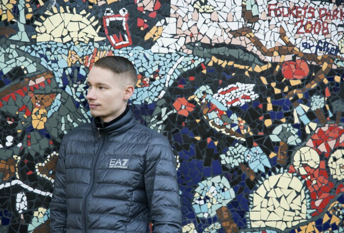 En ung man i täckjacka blickar ut i fjärran, ståendes framför en mosaikvägg.
