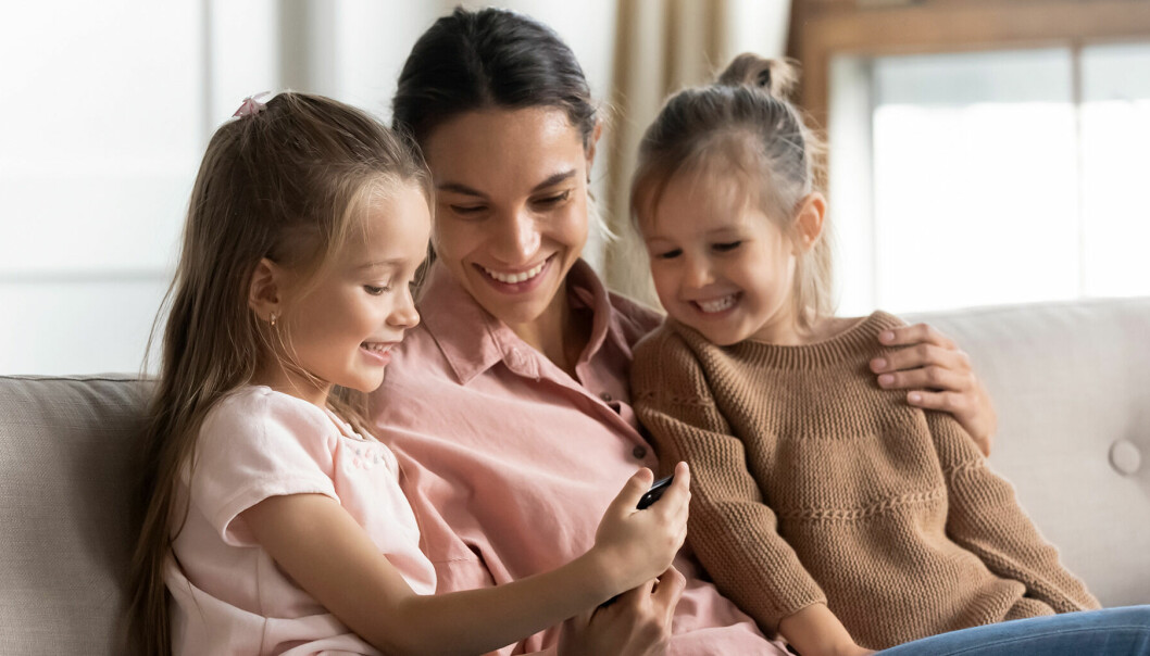 Vuxen kvinna sitter mellan sina två unga döttrar i en soffa. De kollar på en telefon och skrattar.