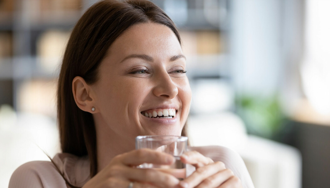 Kvinna i 30-årsålders håller i ett glas vatten och ler.