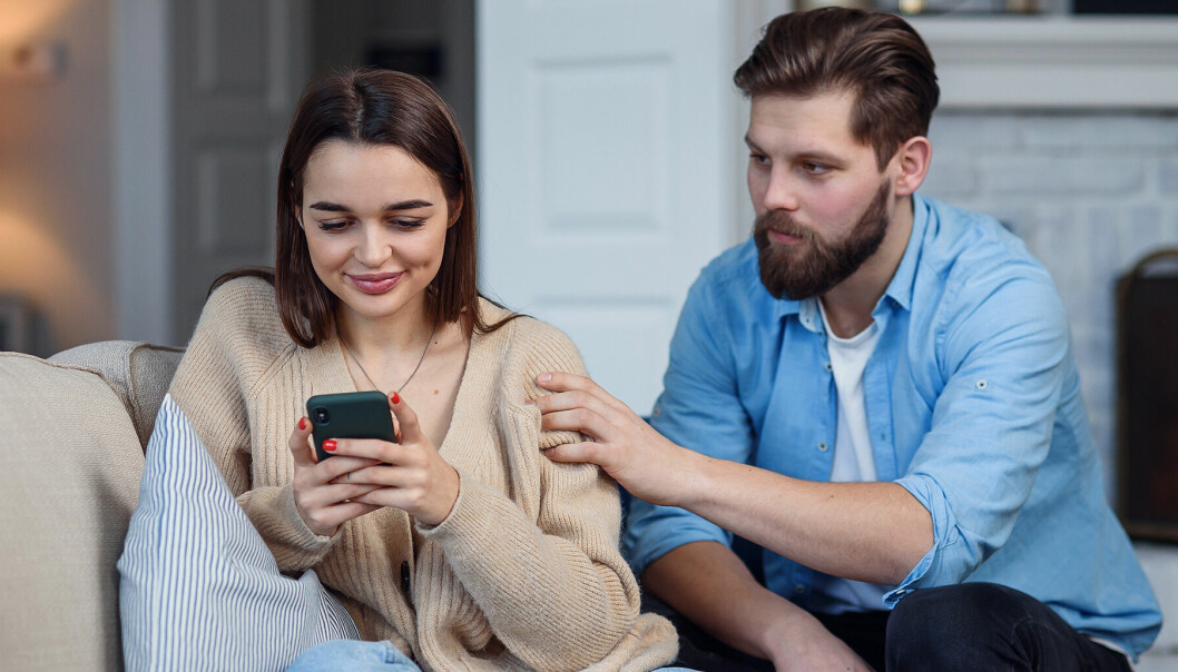 Kvinna i 30-årsåldern sitter i soffan och kollar ner i sin mobiltelefon. Bredvid henne sitter en man i samma ålder och försöker få hennes uppmärksamhet.