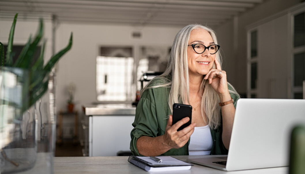 Kvinna sitter vid datorn med telefonen i handen och surfar.