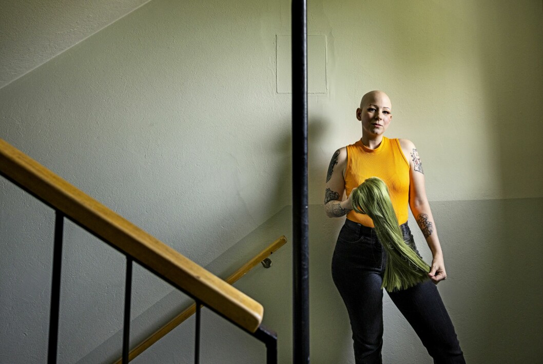 Som tonåring ville hon inte att folk skulle veta, men numera känns det helt naturligt för Lena att inte ha hår. Lena Jakobsson har alopeci och kal hjässa. Hon står i trapphuset med en grön, långhårig peruk i händerna.