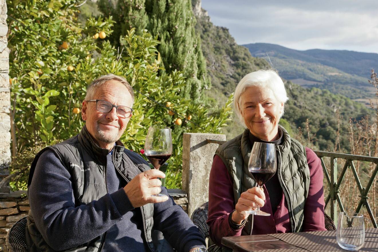 Gunnar och Lotta sitter på terassen till sitt pensionat i franska byn Vieussan, håller var sitt glas rött vin i handen och i bakgrunden syns sluttningen som byn ligger på och berg.