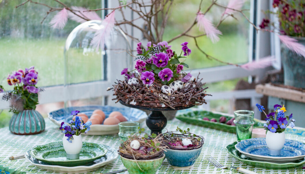 Dukat påskmiddagsbord med blomsterarrangemang i lila toner skapat kring ett fat på fot.