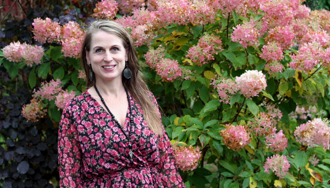 Paula står framför en stor blombuske med rosa blommor. Hon har på sig en blommig klänning.