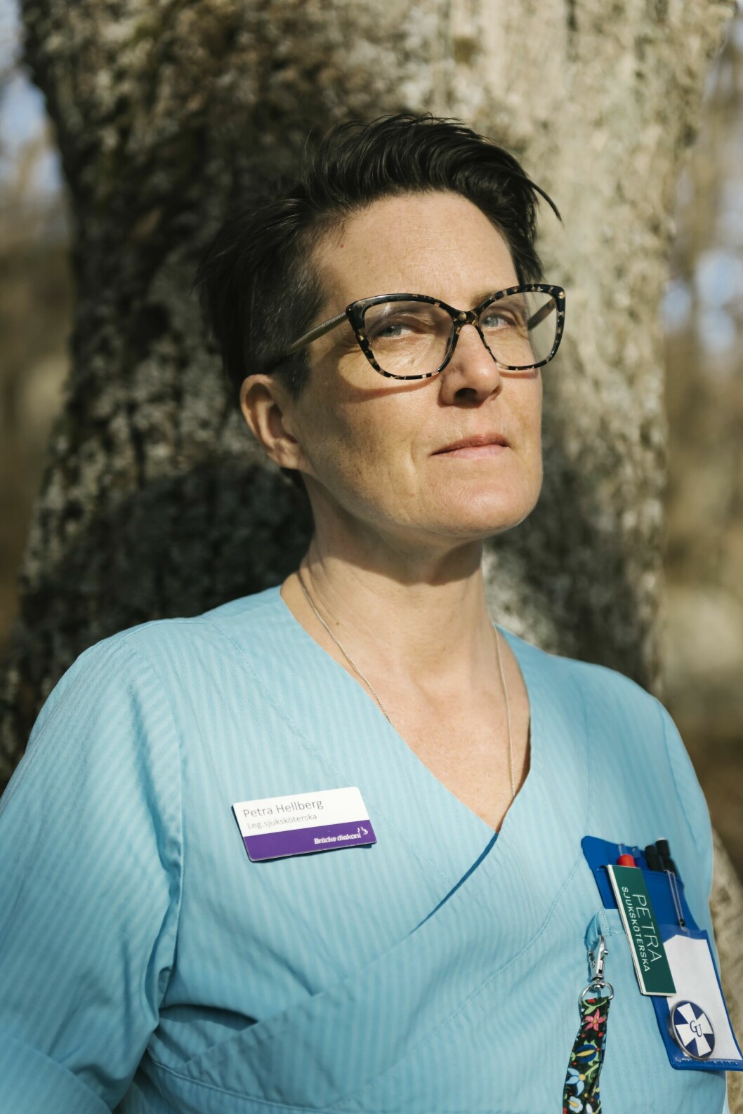 Petra Hellberg står utomhus, lutad mot ett träd, iförd sina arbetskläder med namnskylt, och ser allvarlig ut.
