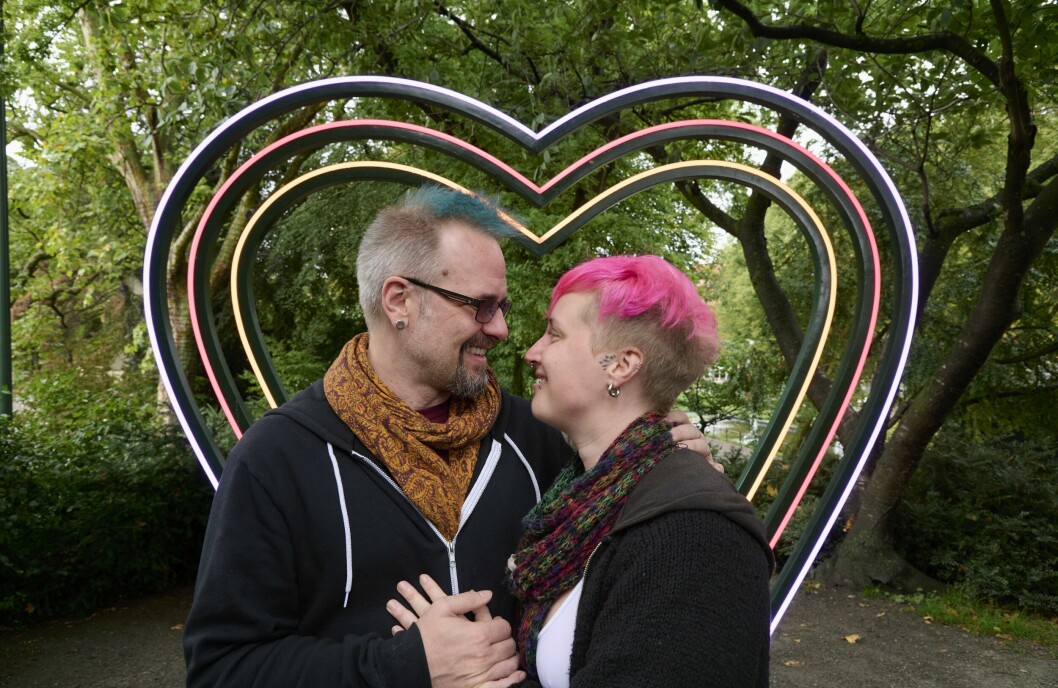 Polyamorösa paret Patrik och Anna står framför en hjärtskulptur. De tittar varandra djupt i ögonen och ler.