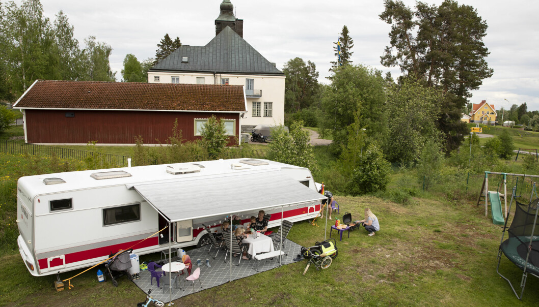 Att ge sig ut på vägarna och alltid ha sitt hem med sig ger familjen Fridland-Engström en känsla av stor frihet. Drönarbild på familjen Fridland-Engströms stora husvagn som står parkerad på familjens tomt. Utanför husvagnen grillar Linda korv och Erik sitter med barnen runt middagsbordet.