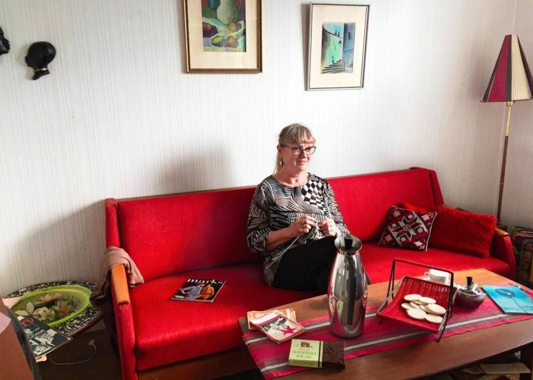 Känslan i ”Andra våningen” ska vara att man hälsar på hos en 60-talsfamilj som bara är ute en liten stund, menar Monica Galle, en av lägenhetens skapare. Foto: Ann-Christin Mathiasson Monica Galle sitter i en röd soffa och stickar i en lägenhet inredd i 1960-talsstil.