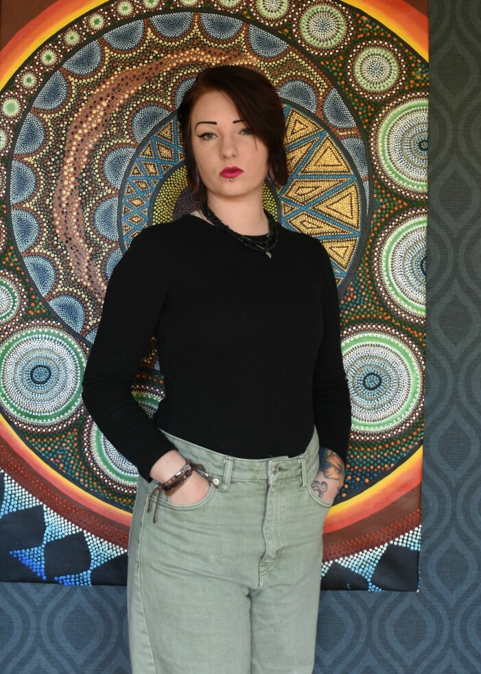 I dag har Sandra en stor passion för konst. Tavlan i bakgrunden har hon själv målat under sina konststudier. Sandra Lundberg framför en tavla med ett detaljerad mönster likt en mandala.