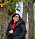 Sara har på sig en svart jacka och röd blus. Hon står utomhus framför ett träd.
