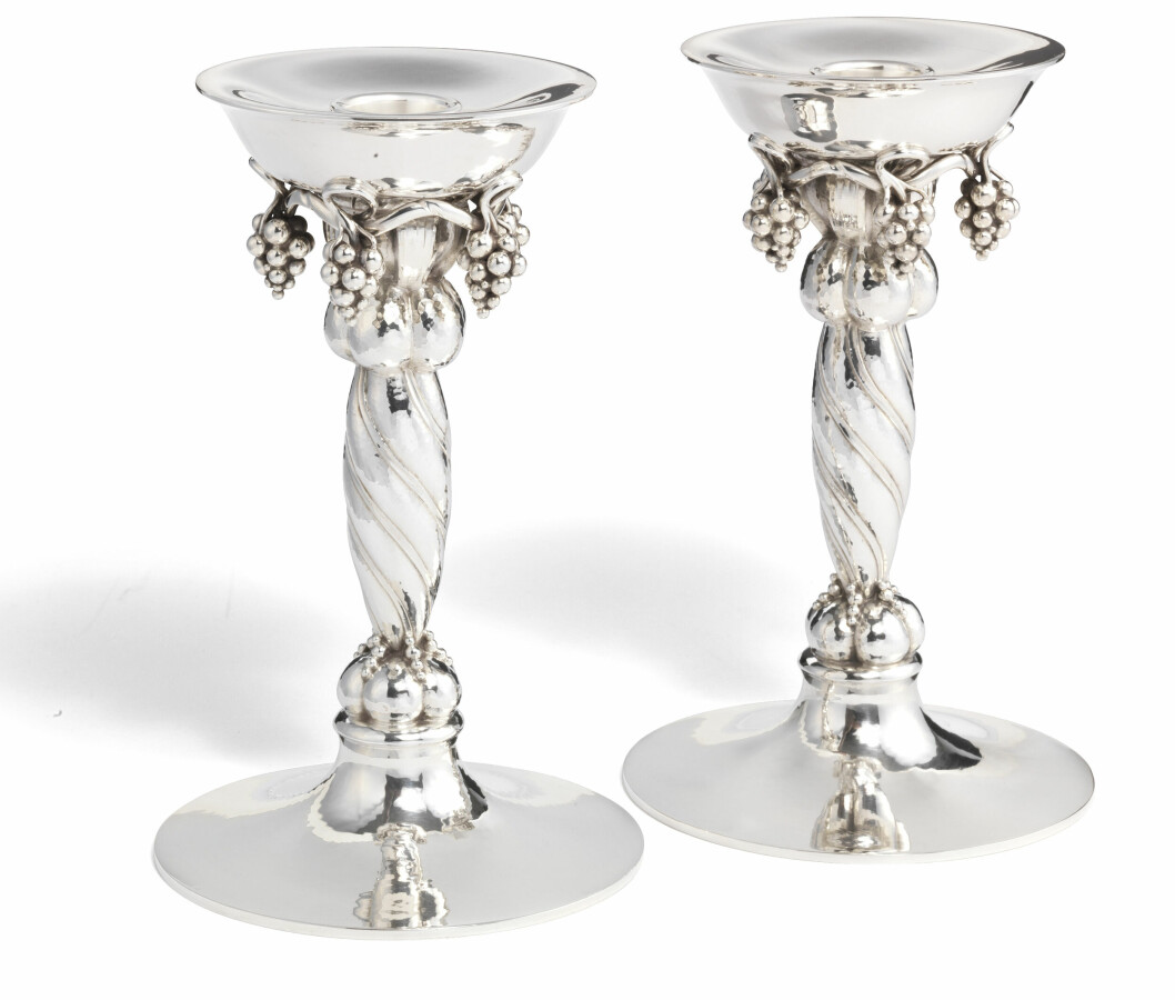 De läckra silverljusstakarna är tillverkade i en modell med vridet skaft och hängande druvklasar som är formgiven av Georg Jensen (1866–1935) 1918. Auktionens par, som är tillverkade under 1900-talets tredje kvartal, såldes 2019 genom Bruun Rasmussen som redovisade ett klubbslag på 50000 danska kronor. Två silverljusstakarna med vridet skaft och hängande druvklasar.