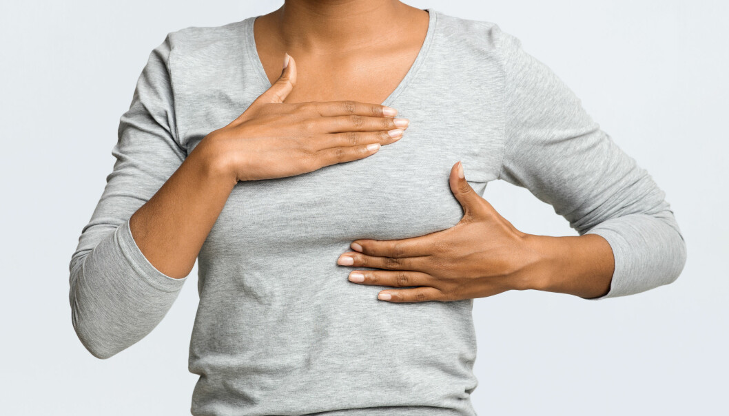 Kvinna i grå långärmad tröja känner på sitt vänstra bröst med båda händerna.