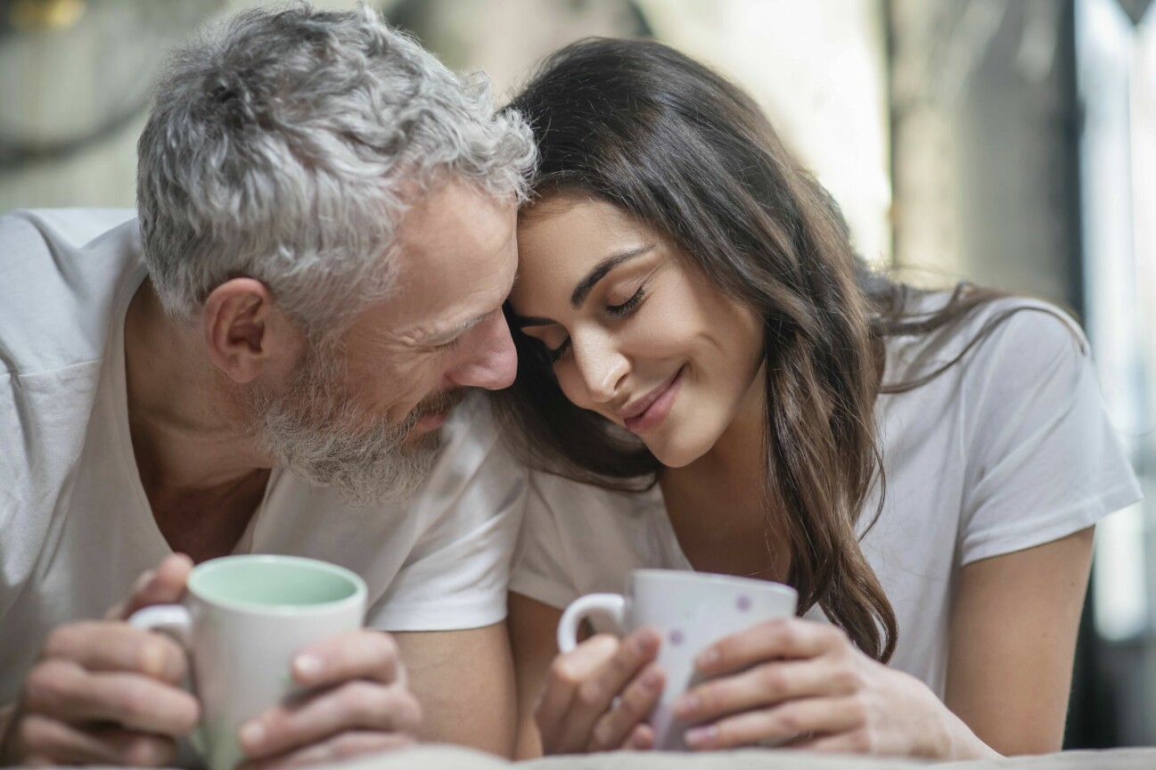 Man och kvinnan sitter tätt bredvid varandra, de ler mot varandra och håller i varsin kaffekopp.