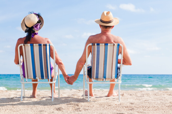 Äldre par som sitter i varsin solstol på stranden och håller hand.