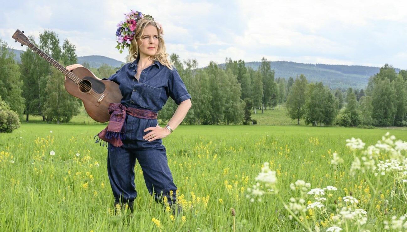 Artisten Sofia Karlsson på en sommaräng med gitarren på höften och blommor i håret.