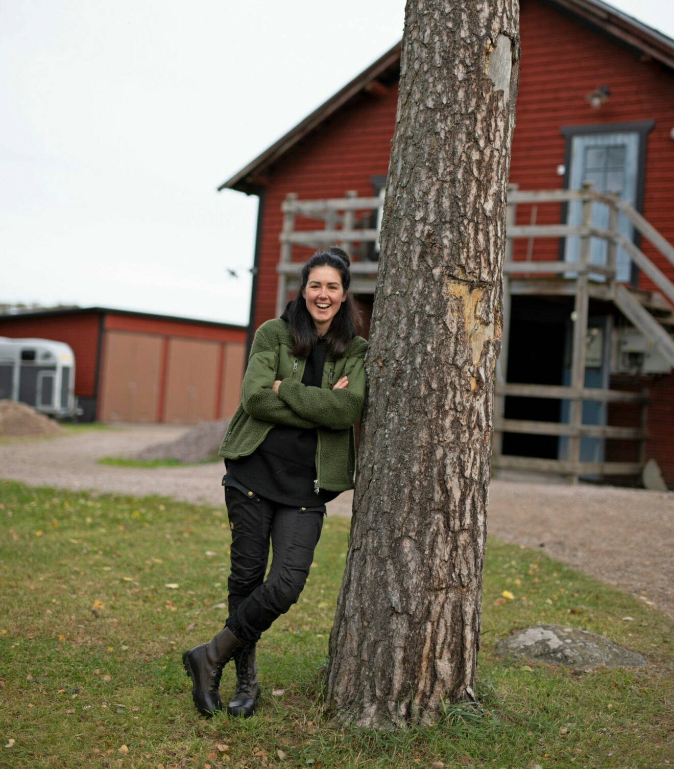 Sofie skrattar och lutar sig mot träd framför en röd byggnad.