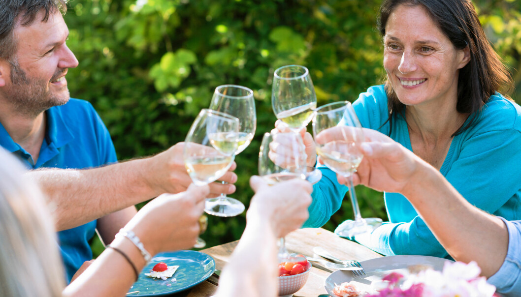 Vänner skålar utomhus med vitt vin i glasen. En medelålders man tittar intresserat på kvinnan intill.