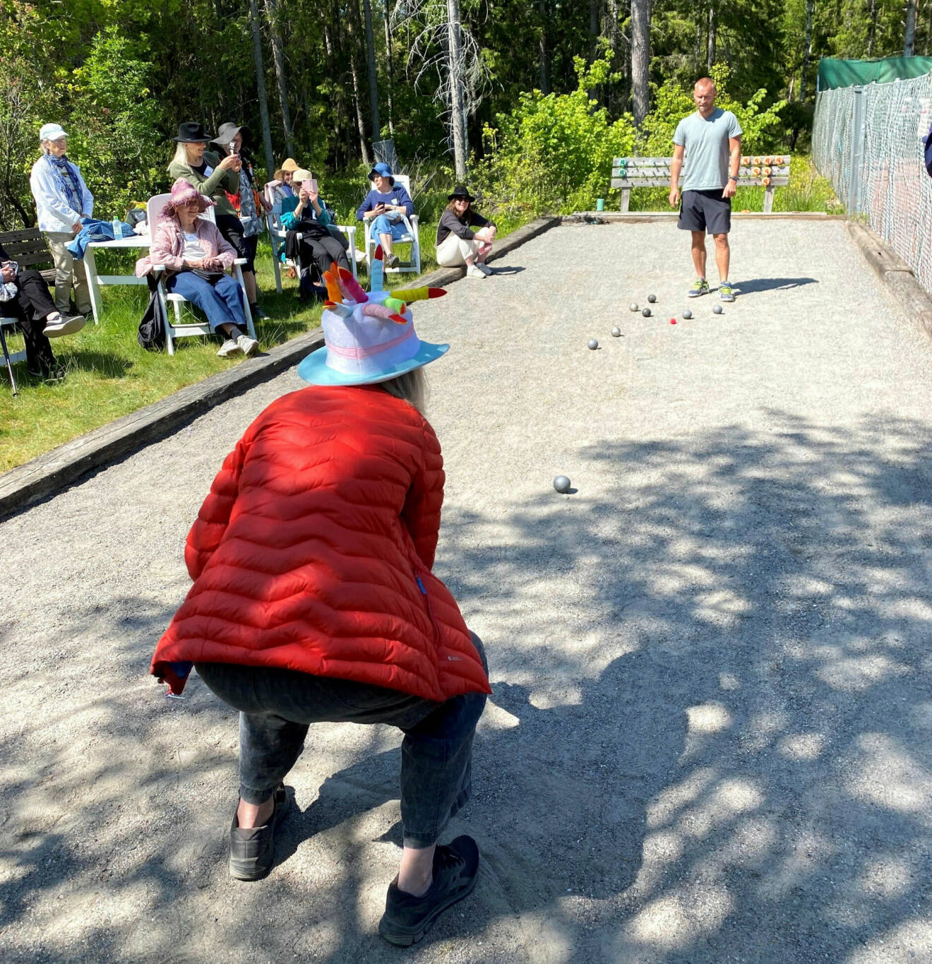 Kvinna med hatt och röd jacka spelar boule på grusplan med man i shorts och t-shirt. Personer, också med hattar, sitter i solstolar intill och tittar på.