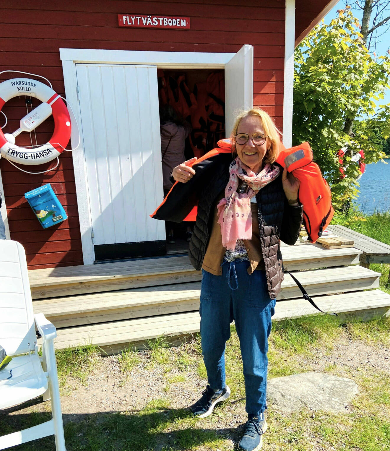 Kvinna med jeans, glasögon och sneakers tar på sig en flytväst ute vid trappan till ett litet rött hus med skylt där det står "Flytvästboden".