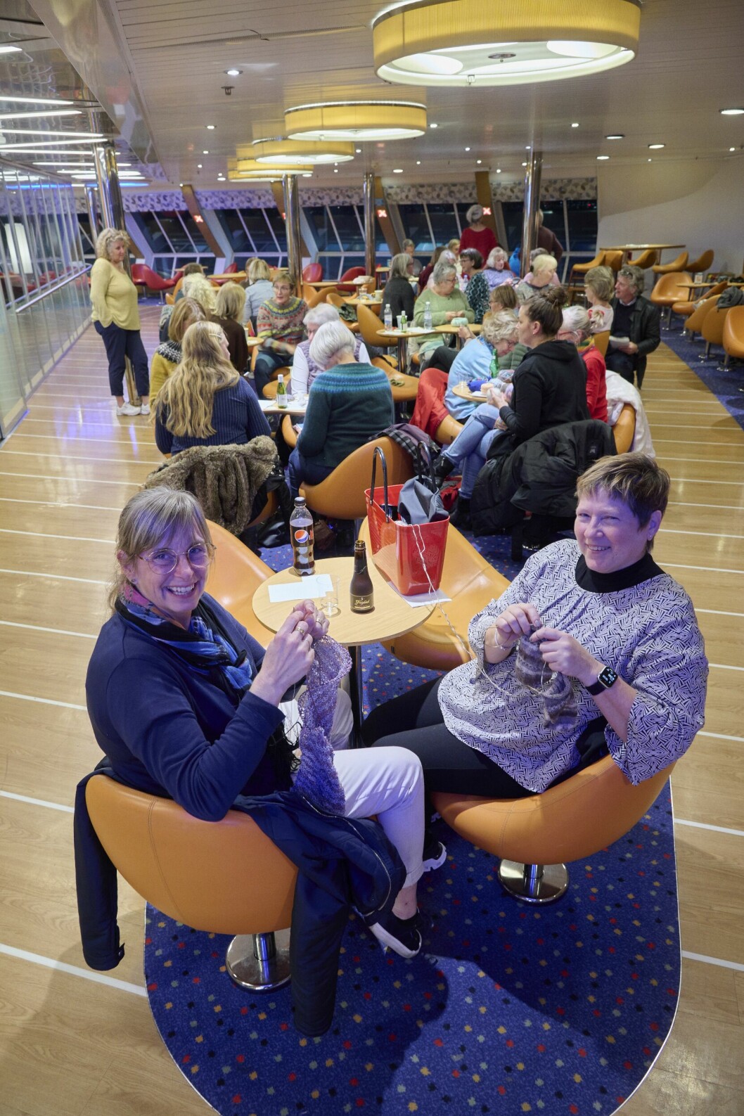 Ska vi sticka och tura? Ja, den här kvällen slöt ett 30-tal upp, däribland Susanne Jönsson och Katarina Roos. Ett 30-tal damer kring borden, de två närmast i bild stickar för fullt samtidigt som de tittar upp och ler. I bakgrunden ses färjefönster ut mot den mörka kvällen.