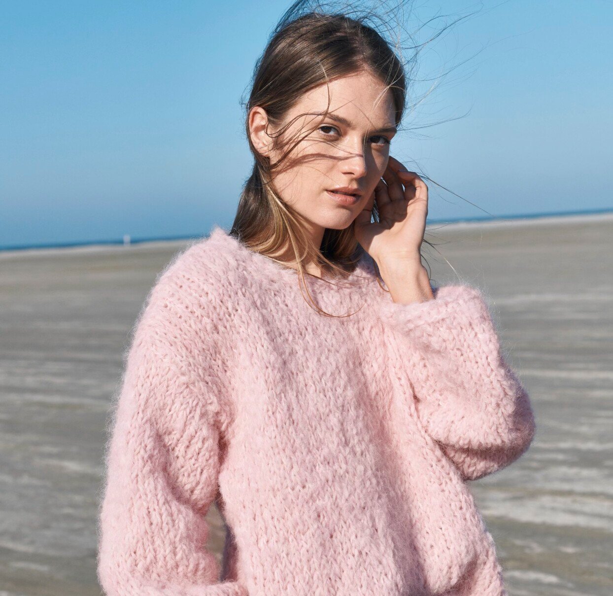Foto: Lana Grossa Kvinna i den fluffiga rosa tröjan på en blåsig strand.