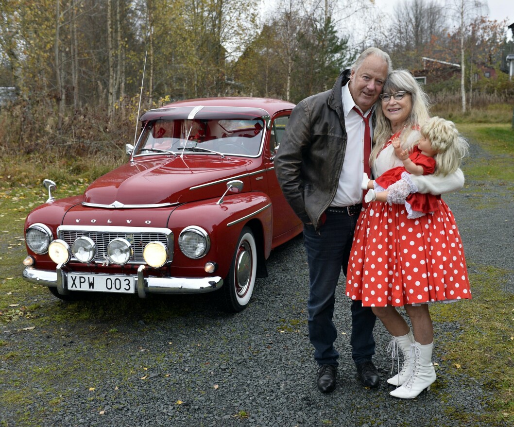 Sune och Lillian Pettersson framför sin röda Volvo PV. De bär tidsenliga kläder. Lillian är iklädd en röd klänning med vita prickar och har en av sina samlardockor under armen.